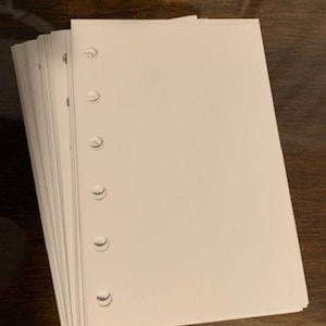 以下が含まれることがあります： オフホワイトの穴あき紙の束。各シートには、左端に沿って6つの等間隔の穴があります。紙は濃い茶色の木製の表面に積み重ねられています。