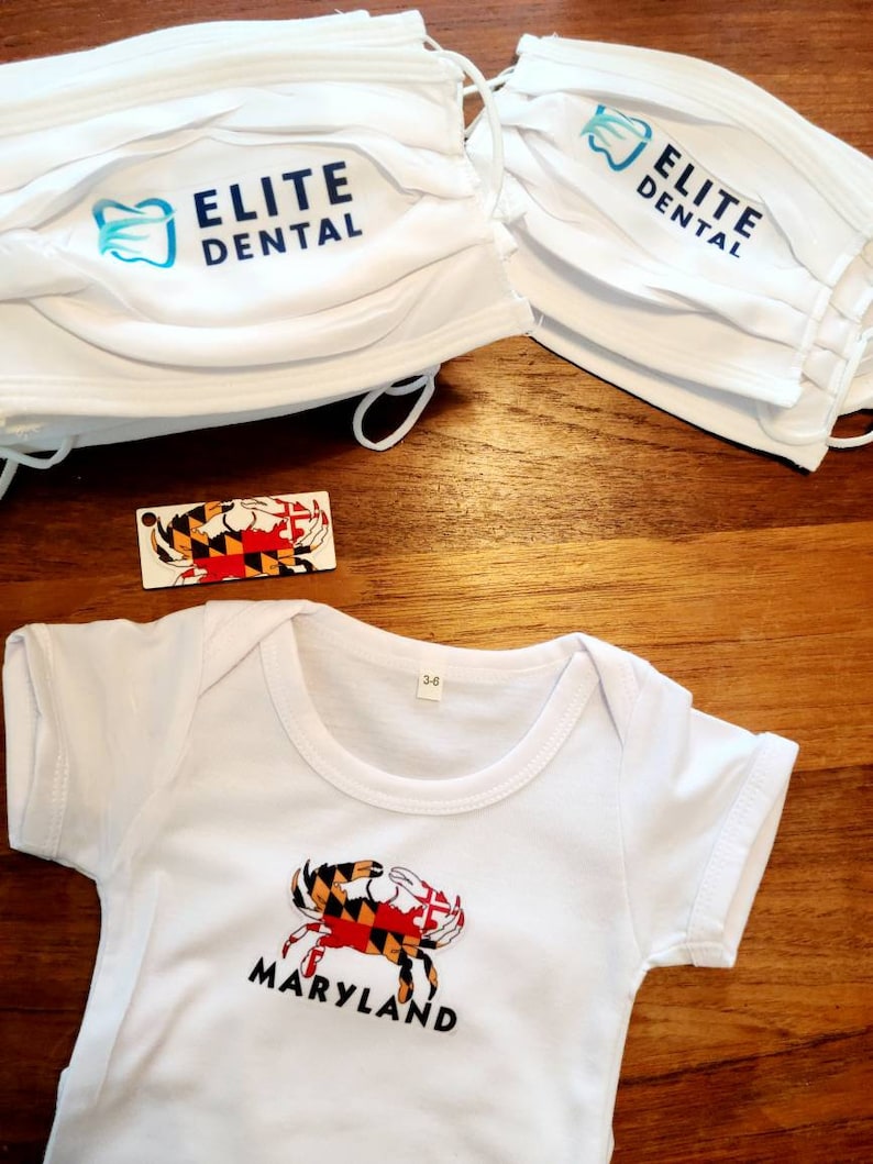 K&ouml;nnte beinhalten: Ein wei&szlig;er Baby-Strampler mit einer Maryland-Krabbe und dem Wort "Maryland" auf der Vorderseite. Der Strampler liegt auf einer Holzoberfl&auml;che mit zwei wei&szlig;en Gesichtsmasken mit dem Aufdruck "Elite Dental" und einer kleinen Maryland-Flagge.