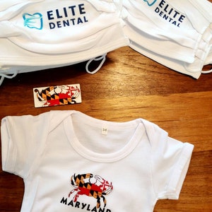 May include: White baby onesie with a Maryland crab and the word "Maryland" printed on the front. The onesie is on a wooden surface with two white face masks with the text "Elite Dental" printed on them and a small Maryland flag.