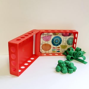 May include: A red plastic storage box with a removable lid. The lid has a colorful floral pattern with circles and lines. The box has multiple round holes on the sides. There are green plastic pieces in the background.