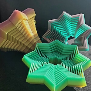May include: Three 3D printed geometric shapes, one is a multi-layered starburst in shades of green, another is a multi-layered starburst in shades of purple, and the last is a multi-layered pyramid in shades of yellow and orange.