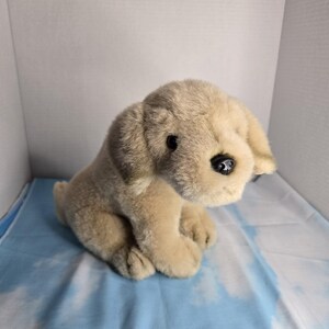 May include: A plush dog toy featuring soft, light tan fur with darker brown shading on the ears. The stuffed animal has a rounded head, floppy ears, a small black plastic nose, and dark, round eyes, depicted in a seated, forward-facing position.