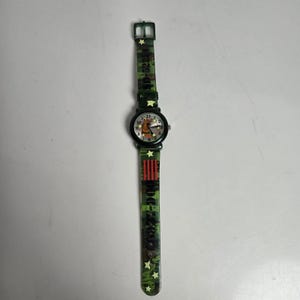 May include: A wristwatch with a round black case and a camouflage-patterned strap. The watch face features a cartoon dog graphic, numbers, and star accents. The strap includes yellow stars, red stripes, and text printed on the camouflage surface.