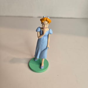 May include: A figurine of a woman in a light blue dress with short sleeves, standing on a green circular base. The woman has orange hair and is wearing purple shoes. The figurine is a collectible item.