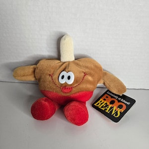 Puede incluir: Un peluche con forma de paleta sonriente y antropomórfica. El juguete tiene un cuerpo marrón, pies rojos y un palo de color claro. La cara presenta ojos grandes y blancos y una sonrisa roja. Una etiqueta dice "BOO BEANS".