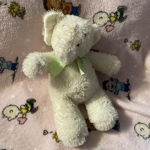 May include: A plush cream-colored elephant stuffed animal with a textured, fuzzy surface. The elephant features large ears, a trunk, and a light green ribbon tied in a bow around its neck. The toy has simple stitched eyes and a soft, rounded body.