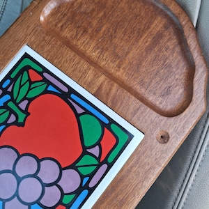 May include: Wooden serving tray with a recessed handle area and a decorative tile insert. The tile features a colorful fruit design with red, purple, green, and blue accents. The tray is made of wood.