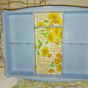 May include: A light blue wall-mounted shelf with a decorative floral print. The shelf has a rectangular shape with a central compartment and several small slots. The floral print features yellow flowers and green leaves.