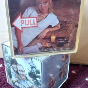 Puede incluir: Un cubo de fotos de plástico transparente con imágenes en los lados. La sección superior presenta a una mujer con un vestido blanco con la palabra "PULL" en una flecha. La sección inferior muestra una pareja y otras fotos.