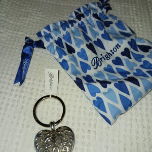 Może przedstawiać: Srebrny brelok do kluczy w kształcie serca z misternym wzorem filigranu. Zawiera niebiesko-białą torbę ze sznurkiem w kształcie serca ze słowem "Brighton" i etykietę z tą samą nazwą marki.