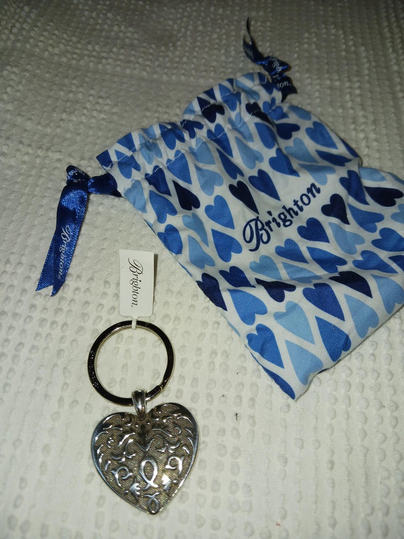 Może przedstawiać: Srebrny brelok do kluczy w kształcie serca z misternym wzorem filigranu. Zawiera niebiesko-białą torbę ze sznurkiem w kształcie serca ze słowem "Brighton" i etykietę z tą samą nazwą marki.