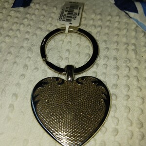 Może przedstawiać: Srebrny brelok do kluczy w kształcie serca z teksturowaną powierzchnią. Brelok ma czarny pierścień i małą dołączoną metkę. Serce ma dekoracyjne akcenty wzdłuż krawędzi.