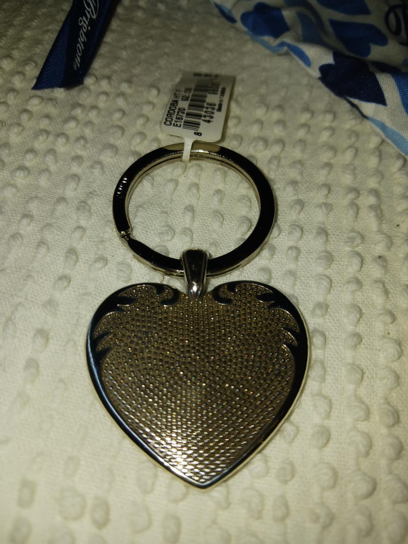 Może przedstawiać: Srebrny brelok do kluczy w kształcie serca z teksturowaną powierzchnią. Brelok ma czarny pierścień i małą dołączoną metkę. Serce ma dekoracyjne akcenty wzdłuż krawędzi.