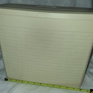 May include: A rectangular, off-white plastic storage bin with a lid and a moulded brick-patterned exterior. A yellow measuring tape is positioned along the base, indicating a width of approximately 30.5 cm for this functional household container.