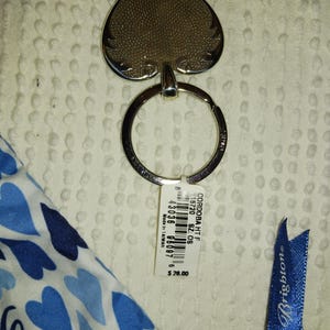 Może przedstawiać: Srebrny brelok do kluczy z zawieszką w kształcie pika. Zawieszka ma teksturowaną powierzchnię i ozdobną obw&oacute;dkę. Widoczna jest r&oacute;wnież etykieta z tekstem oraz niebieska wstążka z napisem "Brighton".