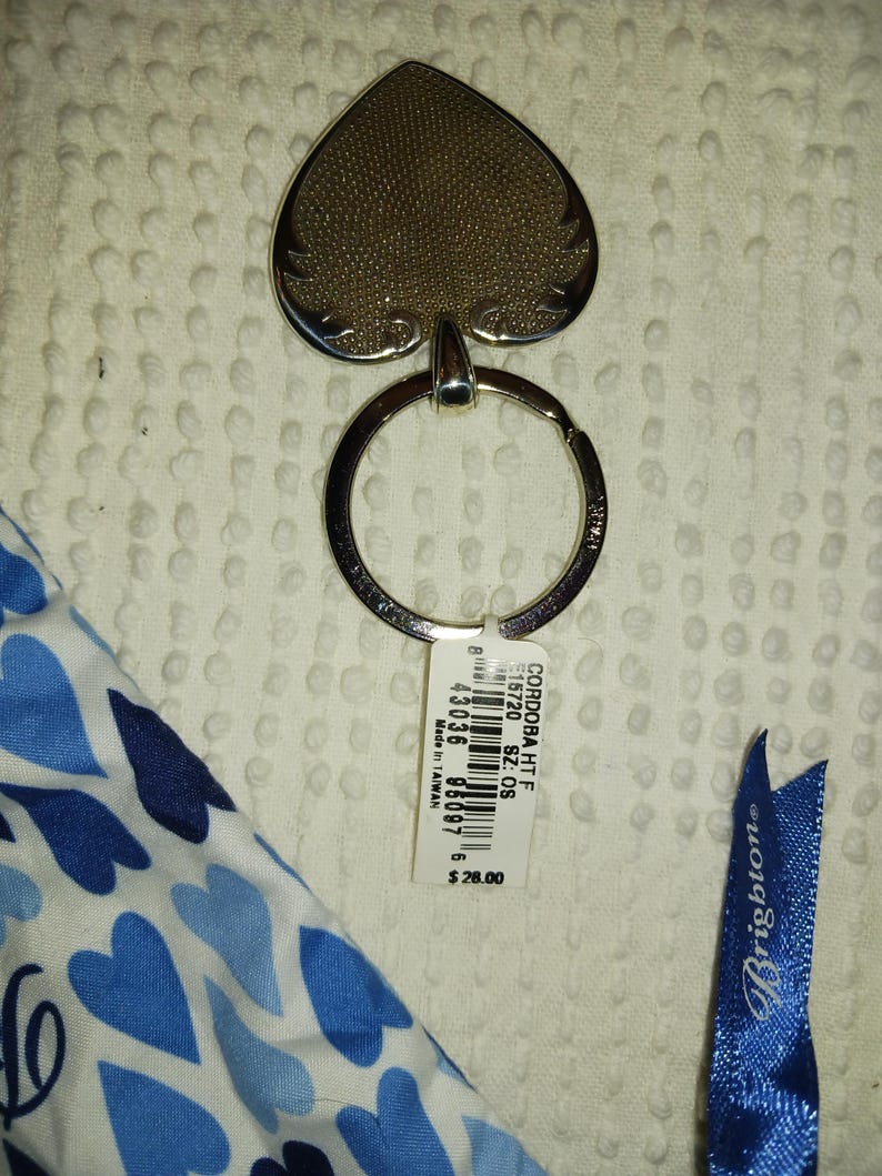 Może przedstawiać: Srebrny brelok do kluczy z zawieszką w kształcie pika. Zawieszka ma teksturowaną powierzchnię i ozdobną obw&oacute;dkę. Widoczna jest r&oacute;wnież etykieta z tekstem oraz niebieska wstążka z napisem "Brighton".