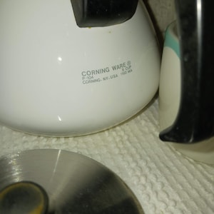 Könnte beinhalten: Eine weiße Corning Ware 6-Tassen-Kaffeekanne mit schwarzem Griff und Edelstahl-Deckel. Der Text "CORNING WARE" und weitere Details sind auf der Seite der Kanne aufgedruckt. Der Deckel ist teilweise geöffnet.