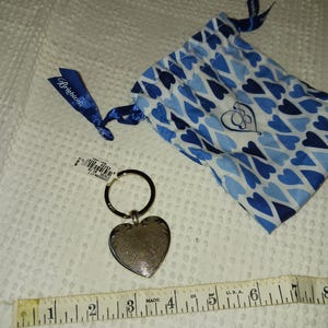 Może przedstawiać: Srebrny brelok do kluczy w kształcie serca z teksturowaną powierzchnią, przymocowany do metalowego k&oacute;łka. Obok breloka znajduje się niebieski woreczek ze sznurkiem i wzorem serca. Brelok ma około 5 cm długości.