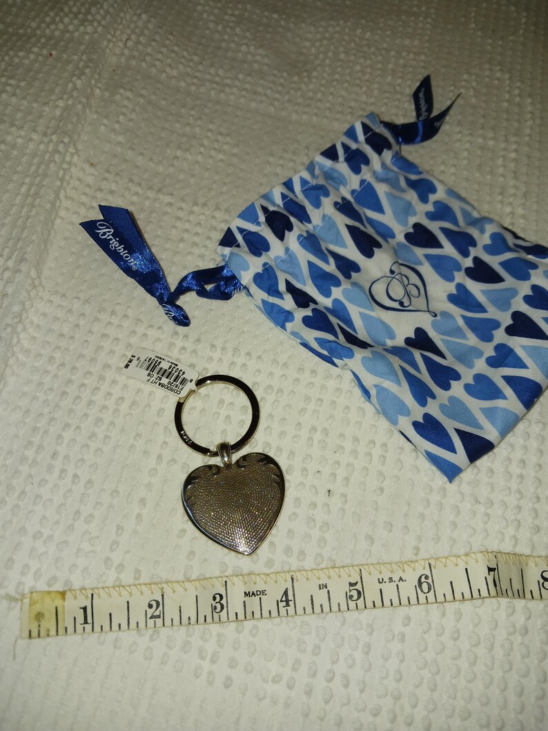 Może przedstawiać: Srebrny brelok do kluczy w kształcie serca z teksturowaną powierzchnią, przymocowany do metalowego k&oacute;łka. Obok breloka znajduje się niebieski woreczek ze sznurkiem i wzorem serca. Brelok ma około 5 cm długości.