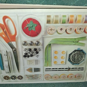 Op de afbeelding: Een naaikit met diverse gereedschappen en benodigdheden. Inclusief oranje schaar, een rode tomaten speldenkussen, spoelen garen, een groene rits, knopen, naalden en een meetlint. De kit is georganiseerd in een witte lade.