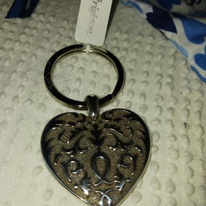 Może przedstawiać: Srebrny brelok do kluczy w kształcie serca z misternym wzorem filigranowym. Brelok ma duży pierścień i małą etykietę z napisem "Brighton". Widoczna jest r&oacute;wnież niebieska wstążka z napisem "Brighton".