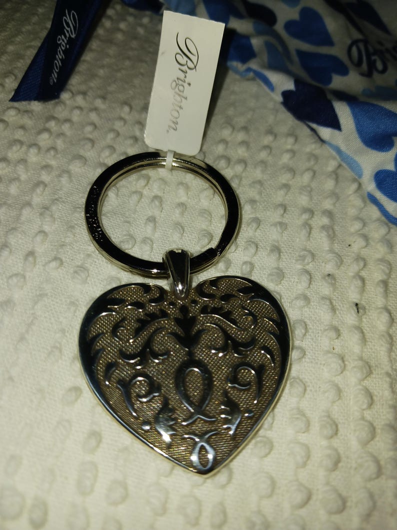 Może przedstawiać: Srebrny brelok do kluczy w kształcie serca z misternym wzorem filigranowym. Brelok ma duży pierścień i małą etykietę z napisem "Brighton". Widoczna jest r&oacute;wnież niebieska wstążka z napisem "Brighton".