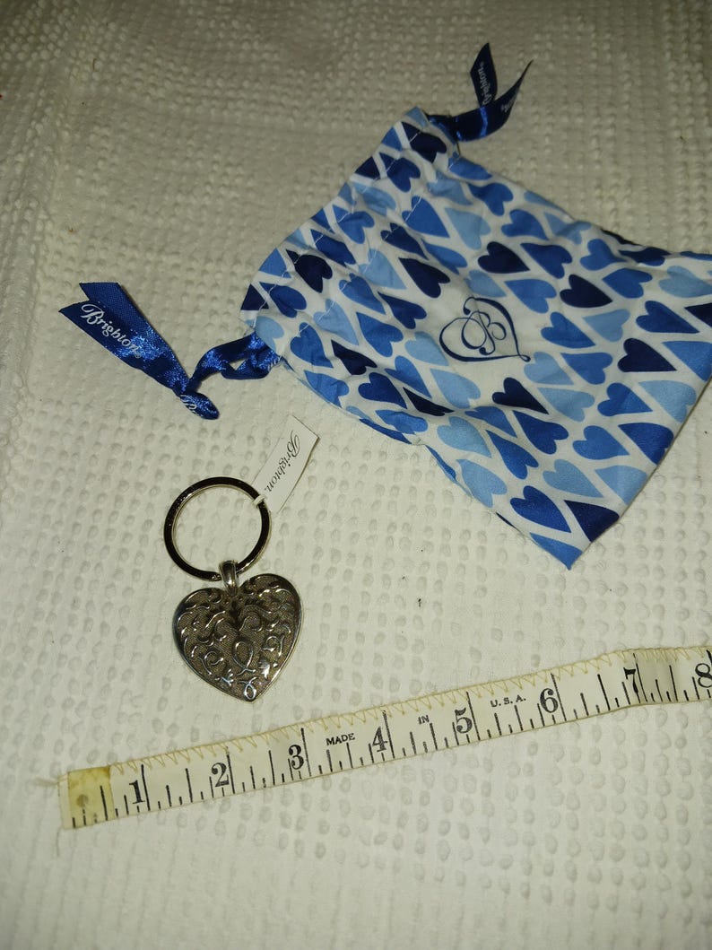 Może przedstawiać: Srebrny brelok do kluczy w kształcie serca z misternym wzorem filigranowym, pasujący niebieski woreczek ze sznurkiem z motywem serca i miarka. Brelok do kluczy znajduje się obok metki z nazwą marki.