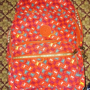 Peut inclure: Sac à dos à roulettes rouge avec un motif répété de figures abstraites blanches, turquoise et jaunes. Le sac à dos a des fermetures éclair dorées, un logo orange rond et des roues noires. Conçu pour le voyage et le transport d'articles.