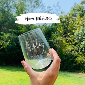 May include: A clear stemless wine glass with the name "Ashley", the title "Bridesmaid", and the date "10-20-2020" printed in white. The glass is held up against a background of green grass and trees.