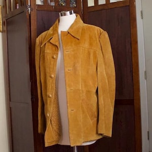 May include: A brown suede jacket with a button-up front and a collar. The jacket is on a mannequin in front of a wooden screen.