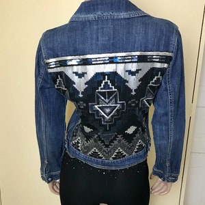 May include: A blue denim jacket with a black and silver sequined design on the back. The design features geometric shapes and patterns.