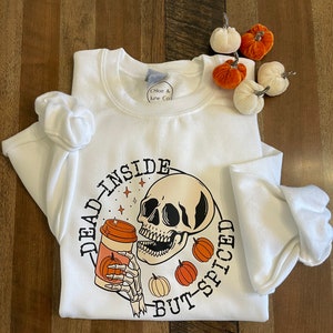 May include: White sweatshirt with a black and white illustration of a skull holding a coffee cup with a pumpkin on it. The text "Dead Inside But-Spiced" is written around the skull.