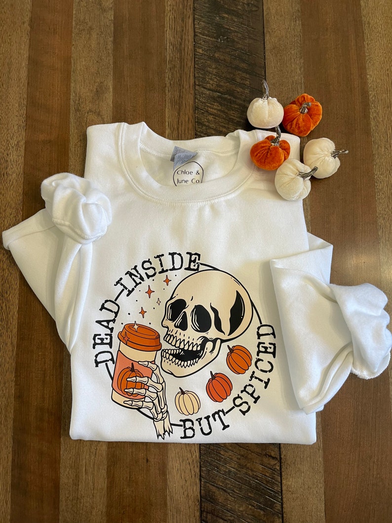 May include: White sweatshirt with a black and white illustration of a skull holding a coffee cup with a pumpkin on it. The text "Dead Inside But-Spiced" is written around the skull.
