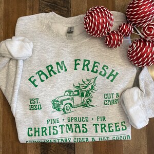 May include: Gray sweatshirt with a green graphic design that says "Farm Fresh" with a vintage truck carrying a Christmas tree. The text also says "Est. 1980", "Pine Spruce Fir", "Christmas Trees", and "Complimentary Cider & Hot Cocoa".