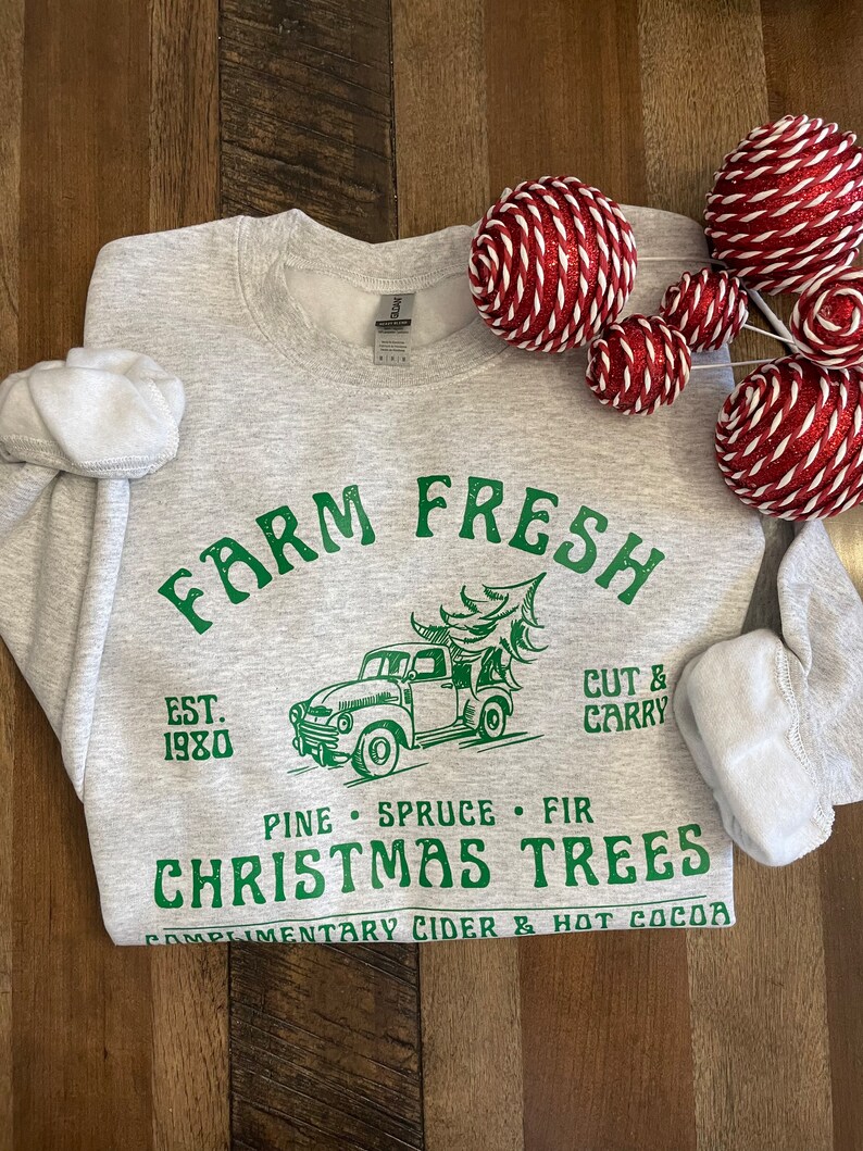 May include: Gray sweatshirt with a green graphic design that says "Farm Fresh" with a vintage truck carrying a Christmas tree. The text also says "Est. 1980", "Pine Spruce Fir", "Christmas Trees", and "Complimentary Cider & Hot Cocoa".