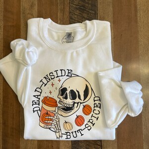May include: White sweatshirt with a graphic of a skull drinking a pumpkin spice latte. The text around the graphic reads "Dead Inside But-Spiced".