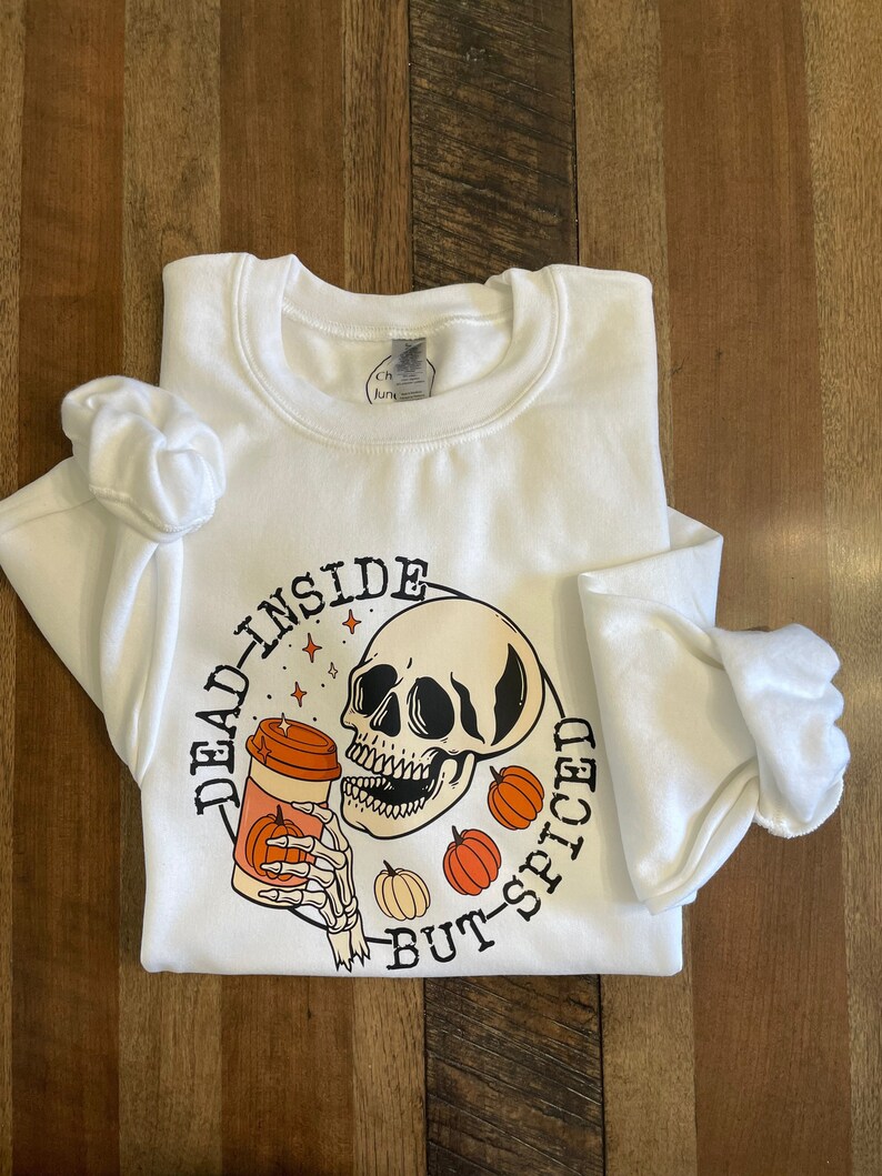 May include: White sweatshirt with a graphic of a skull drinking a pumpkin spice latte. The text around the graphic reads "Dead Inside But-Spiced".