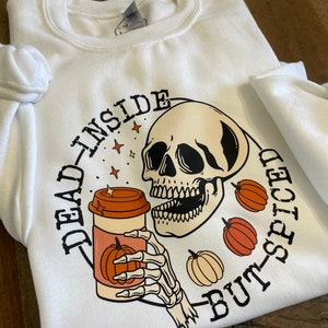 May include: White sweatshirt with a black and orange graphic of a skeleton holding a coffee cup with a pumpkin on it. The text "Dead Inside But-Spiced" is written around the graphic.