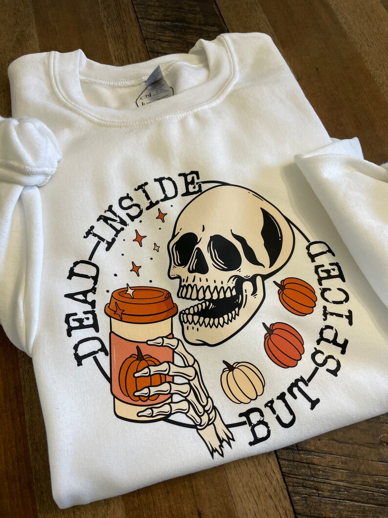 May include: White sweatshirt with a black and orange graphic of a skeleton holding a coffee cup with a pumpkin on it. The text "Dead Inside But-Spiced" is written around the graphic.