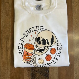 May include: White sweatshirt with a black and white illustration of a skull holding a pumpkin spice latte. The text "Dead Inside But-Spiced" is written around the skull.