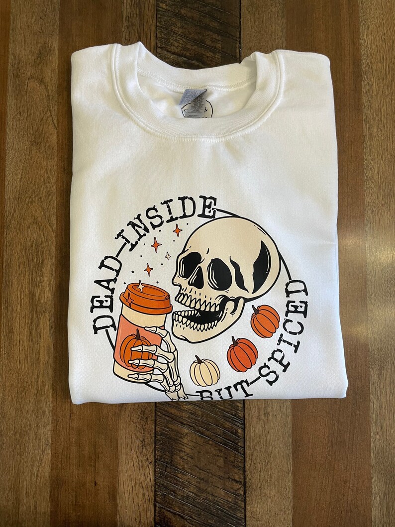 May include: White sweatshirt with a black and white illustration of a skull holding a pumpkin spice latte. The text "Dead Inside But-Spiced" is written around the skull.
