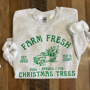 May include: Gray sweatshirt with green text that reads "Farm Fresh Est. 1980 Pine Spruce Fir Christmas Trees" and a green illustration of a pickup truck carrying a Christmas tree. The text "Cut & Carry" is also in green.
