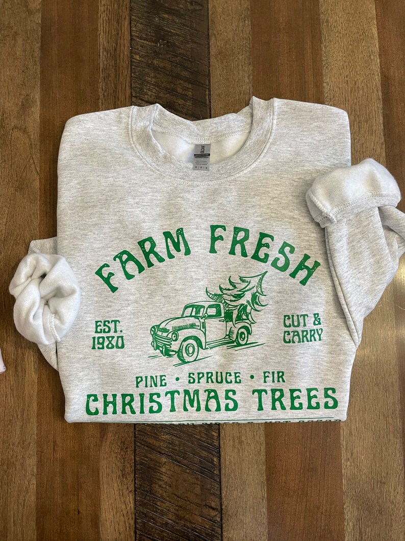 May include: Gray sweatshirt with green text that reads "Farm Fresh Est. 1980 Pine Spruce Fir Christmas Trees" and a green illustration of a pickup truck carrying a Christmas tree. The text "Cut & Carry" is also in green.
