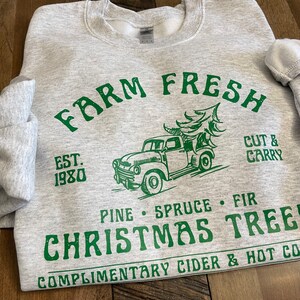 May include: A gray sweatshirt with a green graphic design that says "Farm Fresh" with a vintage truck carrying a Christmas tree. The text also says "Est. 1980", "Cut & Carry", "Pine Spruce Fir", "Christmas Tree", and "Complimentary Cider & Hot Co."