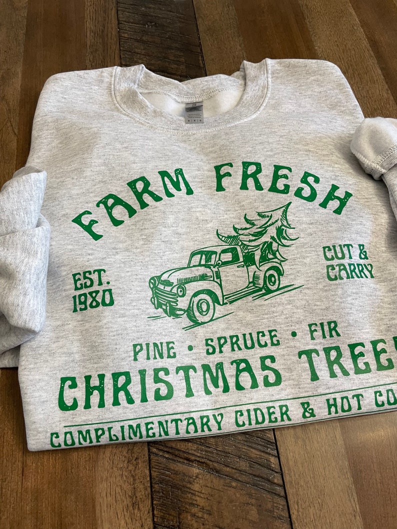 May include: A gray sweatshirt with a green graphic design that says "Farm Fresh" with a vintage truck carrying a Christmas tree. The text also says "Est. 1980", "Cut & Carry", "Pine Spruce Fir", "Christmas Tree", and "Complimentary Cider & Hot Co."