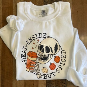 May include: White sweatshirt with a graphic of a skeleton holding a pumpkin spice latte. The text reads "Dead inside but-spiced".
