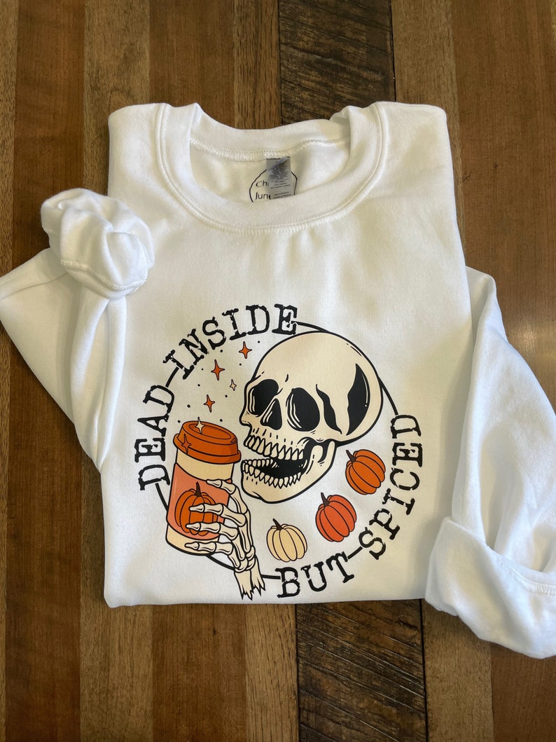 May include: White sweatshirt with a graphic of a skeleton holding a pumpkin spice latte. The text reads "Dead inside but-spiced".