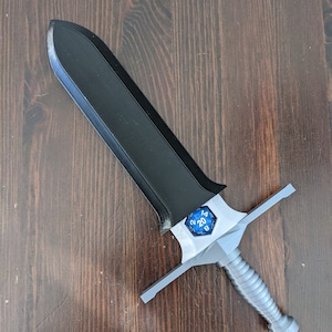 May include: A black and silver sword with a blue 20-sided die on the hilt. The sword is on a wooden surface.
