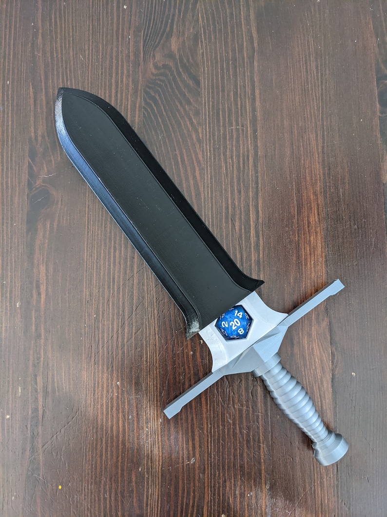 May include: A black and silver sword with a blue 20-sided die on the hilt. The sword is on a wooden surface.