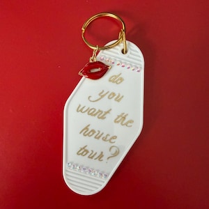 May include: White key tag with gold text that reads "do you want the house tour?" A red lip charm and clear rhinestones adorn the tag, which hangs from a gold key ring. The background is a solid red.